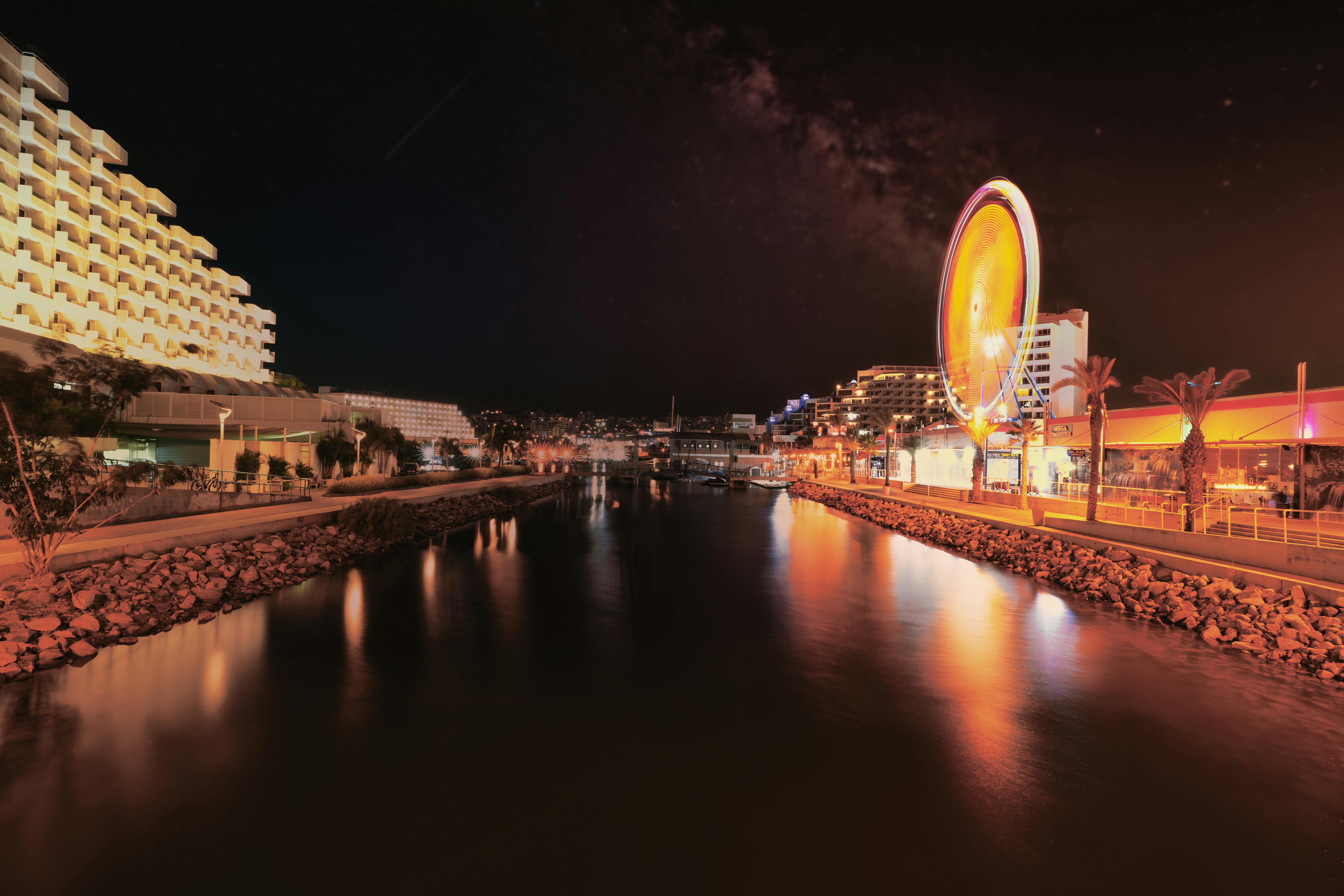 טיילת נופש מוארת בלילה עם גלגל ענק והשתקפויות במים | Illuminated Resort Promenade at Night with Ferris Wheel and Water Reflections