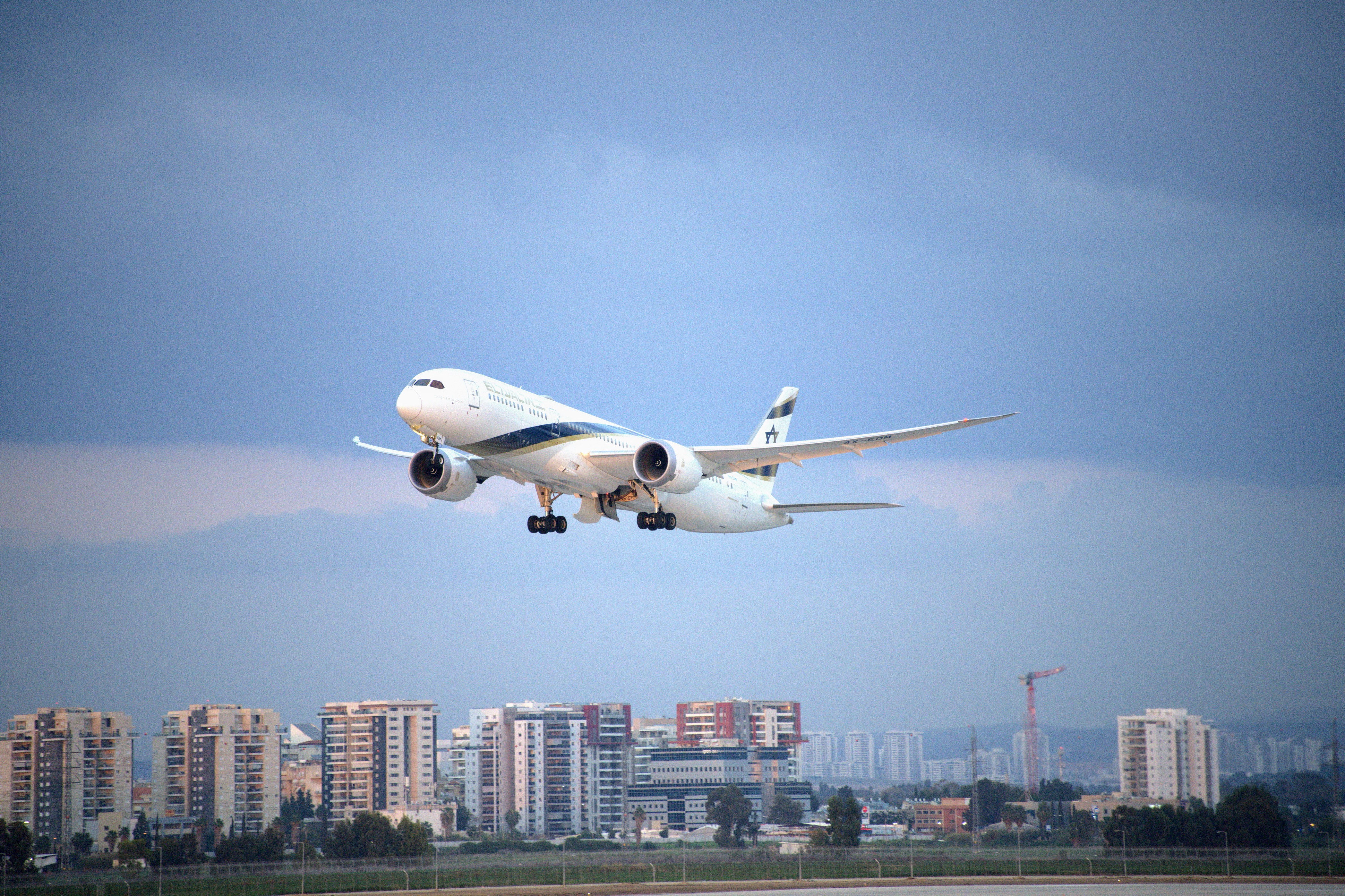 מטוס נוסעים של אל על ממריא על רקע נוף עירוני ושמיים מעוננים | El Al Passenger Airplane Taking Off Against a Cityscape and Cloudy Sky